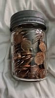 Clear glass jar filled with mostly Canadian pennies and few USA pennies, showing embossed measurement marks on jar and metal lid on top.