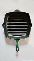 Full view of square Le Creuset cast iron grill pan with green handle and raised grill ridges inside