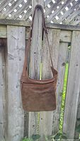 Front view of brown leather crossbody bag showing zipper pocket and strap attached to fence.