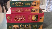 Four game boxes stacked, showing spines: base game, Cities & Knights expansion, two 5-6 player extensions.