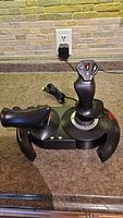 Front view of black Thrustmaster T-Flight Control joystick with integrated throttle and USB cable coiled next to it on a countertop.
