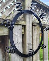 Black circular metal wine rack with eight spiral arms for holding bottles, mounted on wooden fence.