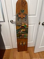 Full view of the hand painted wooden ironing board top standing vertically against white doors on a wooden floor, showing entire painted surface including fruit and farm scenes.
