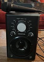 Front view of the Singsation karaoke machine with microphone on top, showing power and control knobs including master volume, music EQ, mic volume, voice FX, and lighting color buttons.