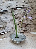 Photo of the small pewter ikebana flower display stand with a floral arrangement inserted into the center hole, textured pewter finish visible, placed against a birch bark background.