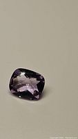 Photo of a cushion cut purple amethyst gemstone with checkerboard facets on a plain background.