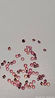 Lot of tiny diamond cut calibrated Padparadscha sapphires scattered on white surface showing orange-pink hues and good clarity.