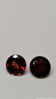 Two round faceted deep red garnet gemstones reflecting light, presented side by side on neutral background.