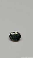 Oval cut green sapphire gemstone shown on neutral background, focused on the polished faceted top surface and visible inclusions.