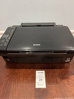 Front and top view of black Epson WiFi printer on wooden floor with visible control panel buttons on left side and brand logo on top center.