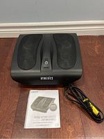 View of the black Homedics foot massager with power button and mesh foot slots along with user manual and power cord on floor.