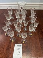 Front view of all 20 assorted cut glassware pieces arranged on floor including an elevated bowl, various stemmed glasses of different sizes with floral and geometric cuts.