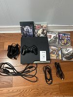 Photo showing PlayStation 3 console with one DualShock 3 controller, six game cases, and several cables on wooden floor.