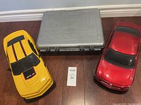 Top view of two 1:43 scale RC cars, one yellow with black and red stripes and one red, alongside a grey plastic storage/transport box.