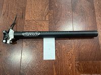 Full side view of the black seat post laid on a wooden floor, showing its length and saddle clamp.