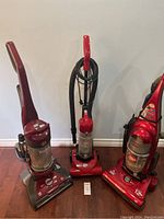 Photo of three red upright vacuum cleaners side by side on a wooden floor against a wall, all showing their front with clear dirt collection canisters visible.