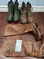 Three pairs of women's boots on wooden floor: tall brown leather boots, green ankle boots, brown lace-up ankle boots