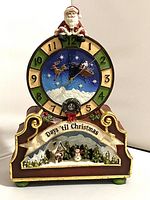Front view of Christmas countdown clock showing Santa at top, 12-numbered clock face with Santa's sleigh in center, and winter village scene at base.