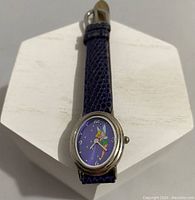 Front view of the oval Disney Timeworks watch with purple dial featuring Tinkerbelle and a purple leather strap.
