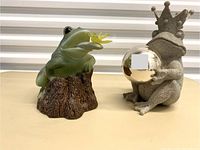 Front view of two playful garden frogs, one green with yellow dragonfly sitting on a tree stump, one gray frog with crown holding mirrored gazing ball