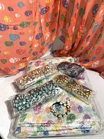 Photo showing 5 packs of individually wrapped skull bead bracelets in various colors with skull-shaped adjustable knots on a multicolored skull pattern scarf and orange skull-patterned fabric background.
