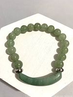 Green jade bracelet laid on a white hexagonal surface showing the round beads and curved jade centerpiece with silver-tone spacers.