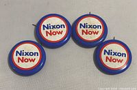 Front view of four Nixon Now 1972 re-election campaign pins with red, white and blue design and visible wear.