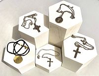 Overview photo showing all five religious necklaces displayed on white geometric platforms, including the black cord necklace with Ave Maria pendant and several silver-tone crosses and medallions.