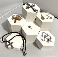 Six religious jewelry items displayed on white hexagonal stands including crosses and a pin