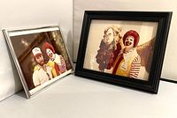 Two framed vintage photos of Ronald McDonald characters, one black framed and one silver framed, shown at side angle.