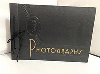 Front angled view of vintage black photo album showing embossed profile and gold lettering 'Photographs' with side ties.