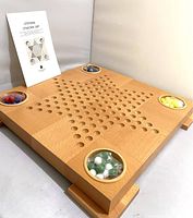 Wooden Chinese Checkers game board with star-shaped indentations and four circular marble holders with different colored marbles. Instruction booklet in the background.