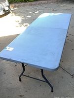 Full view of a light blue rectangular plastic folding table set outdoors on pavement. The table has metal legs and appears lightly used.
