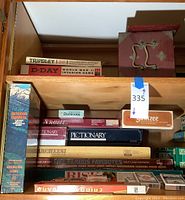Shelf with stacked vintage board games including Tripoley, D-Day World War II game, Scrabble, Pictionary, Dominoes, Outdoor Survival, and Mahjong set box.