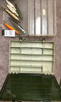 Six large metal fishing lures of varying colors and finishes inside a clear plastic compartment box alongside a green plastic storage box with compartments and a clear lid.