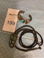 Cuff bracelet and braided leather bolo tie displayed on blue patterned surface next to lot marker 199