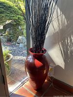 Glossy dark red ceramic floor vase filled with tall black sticks near a window with outdoor garden visible.