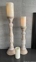 Two carved wooden candle sticks of different heights with three ivory battery operated candles in front, placed on a wooden surface against a grey textured wall.