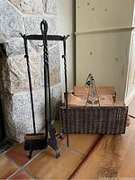 Photo shows black metal fireplace tool set with poker, shovel, and brush on a stand next to a woven wicker basket with firewood, placed on a tiled floor by a stone fireplace.
