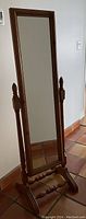 Photo of a tall, rectangular full length mirror with wooden frame and stand, placed indoors on tiled floor against a white wall.