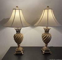 Front view of the pair of decorative table lamps on a black surface against a beige wall, showing both lamps fully assembled, illuminated, with sculpted bases and bell-shaped shades.