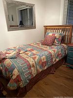 Twin bed made up with quilt, matching pillow shams, bed skirt, and extra decorative pillow. Bedding features red, blue, beige geometric and floral patterns.