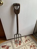 Full view of antique garden fork showing long wooden handle, metal fork tines, and floor background.