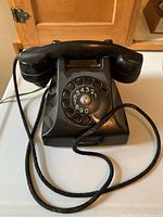 Front view of black rotary dial telephone showing rotary number dial and handset laying on cradle with attached cord.