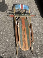 Top-down photo showing the wooden sled, metal runners, steering bar with red handles, and blue padded backrest with stripes and cutout details.