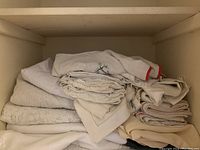 Assorted folded linens stacked in a shelf, shows variety including crochet, embroidered and plain pieces.