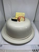 White ceramic covered cake plate with round base and dome lid, featuring cheese-shaped decorative handle on top.