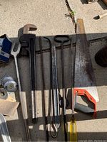 Photo showing large pipe wrench, pry bars, a hand saw with yellow handle and orange frame, and some miscellaneous metal tools on concrete floor.