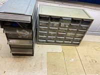 Two gray plastic multi-drawer organizers on a workbench; left organizer has 5 vertical drawers; right organizer has 24 small pull-out drawers