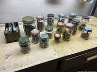Photo of 14 glass jars and 2 metal containers filled with screws, nails, and other fasteners organized on a workbench.